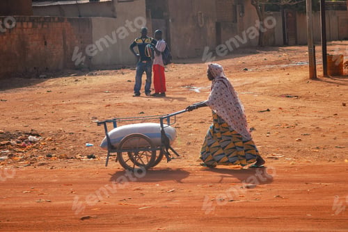 Preview: An old African woman pushing a sack of rice or flour in the sand streets of an African city