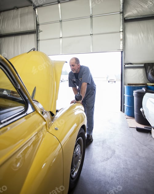 Preview: A mechanic looking under the bonnet of a bright yellow Morris Minor English classic car.