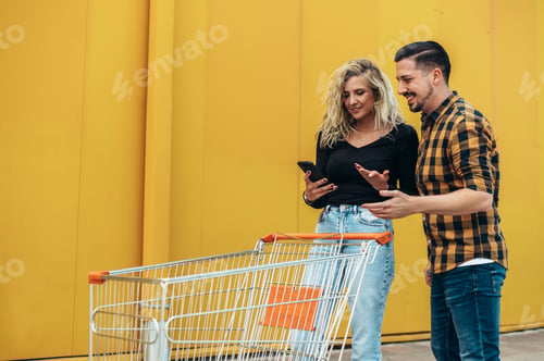 Preview: Young beautiful couple using smartphone while holding a shopping cart