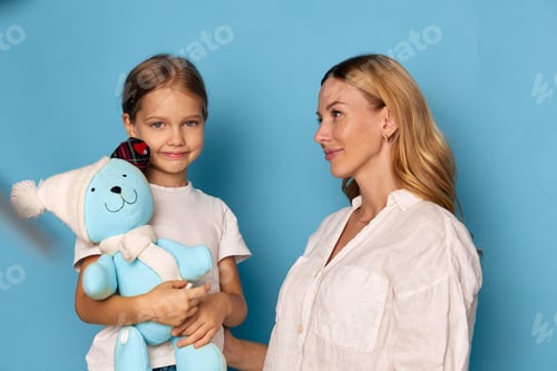Preview: Woman holding blue teddy bear stands beside little girl in white shirt, both smiling and enjoying