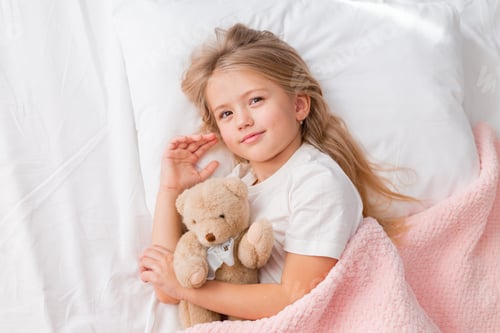 Preview: little blonde girl smiling in a white T-shirt lying in bed under a knitted blanket with a toy teddy