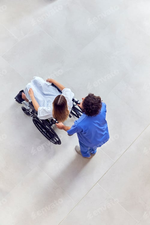 Preview: Overhead Shot Of Nurse Wearing Scrubs Pushing Female Patient In Wheelchair Through Hospital Building