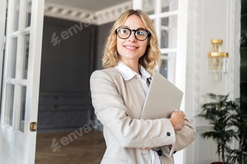 Preview: Smiling businesswoman holding a laptop, standing confidently in an elegant interior.