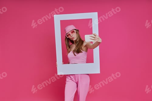 Preview: Playful young woman looking through a picture frame while standing against pink background
