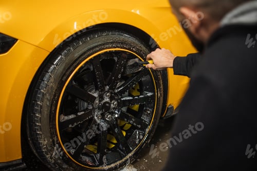 Preview: Close up of handsome young male employee worker cleaning the wheel tire of modern yellow sport car