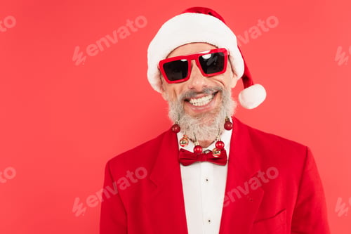 Preview: happy mature man in santa hat and sunglasses with christmas balls on beard isolated on red