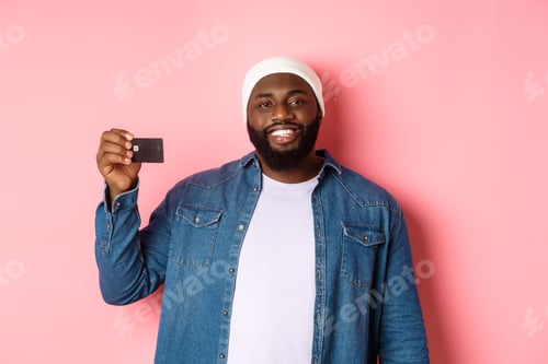 Preview: Shopping concept. Satisfied young bearded man in beanie showing credit card, smiling pleased, making