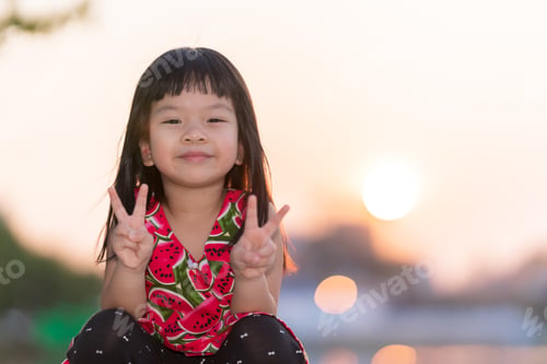 Preview: Girl sitting with sweet smile. Child raised two fingers and look at camera.