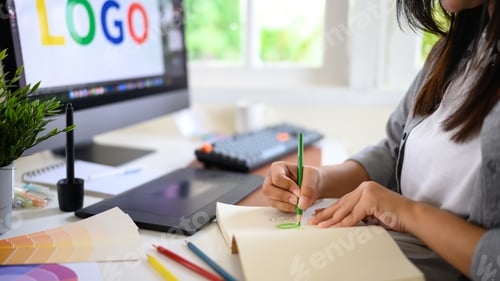 Preview: Closeup creative woman sketching logo ideas in notebook with color samples and pencils