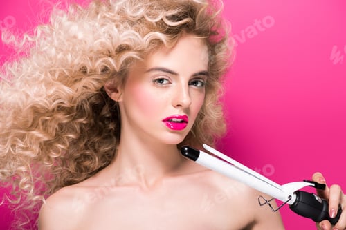 Preview: beautiful naked girl with long curly hair holding hair curler and looking at camera isolated on pink