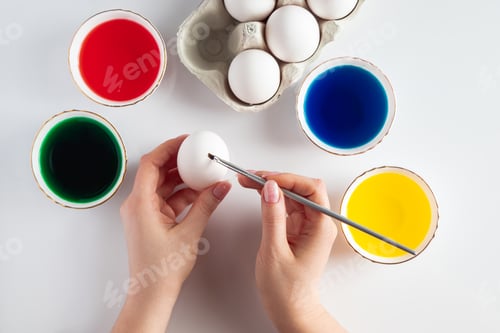 Preview: Young woman draws with colorful paints on white eggs for Easter.