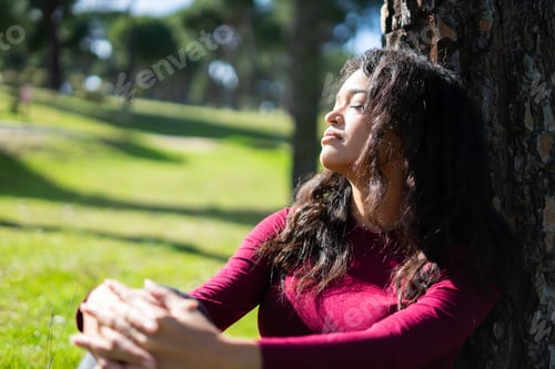 Preview: Latin woman relaxing in park leaning against tree enjoying sunlight