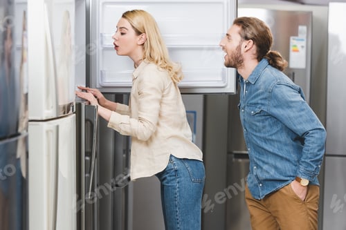 Preview: side view of shocked boyfriend and girlfriend looking at fridge in home appliance store