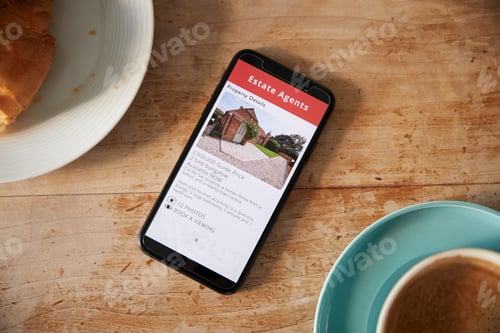 Preview: Close Up Of Mobile Phone With Realtors Property App Lying On Café Table