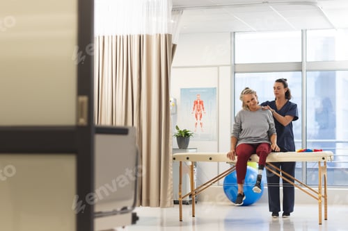 Preview: Caucasian female physiotherapist and senior woman with artificial leg massaging back at hospital