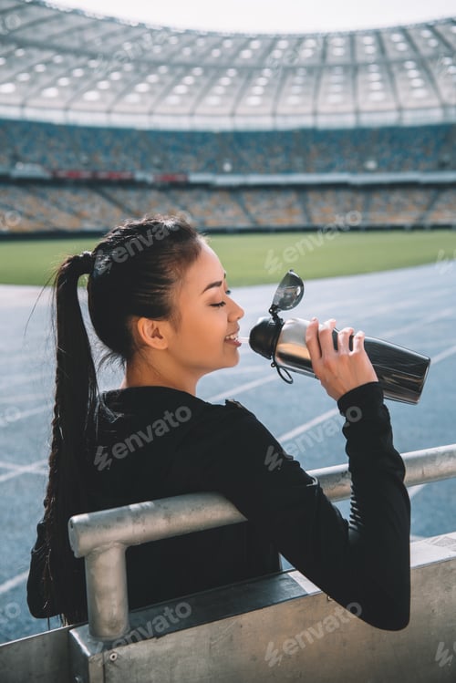 Preview: Smiling young sportswoman drinking water from sports bottle on stadium