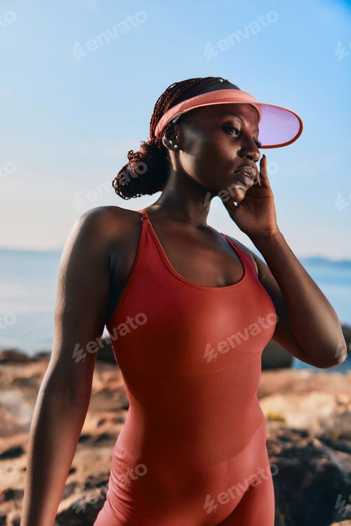 Preview: Woman in a pink visor and red swimsuit posing near the ocean at sunset