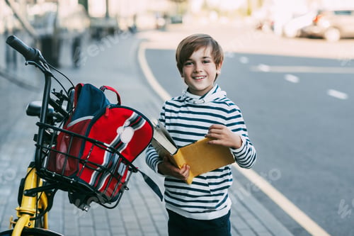 Preview: A cheerful boy holds a book in his hands and stands next to a Bicycle with a backpack hanging