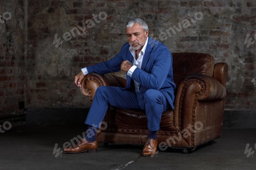 Preview: A man in a blue formal suit sitting on a leather armchair.Portrait of a businessman on a brick wall