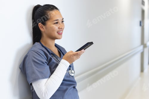 Preview: Biracial female doctor wearing scrubs talking on smartphone in hospital corridor, copy space