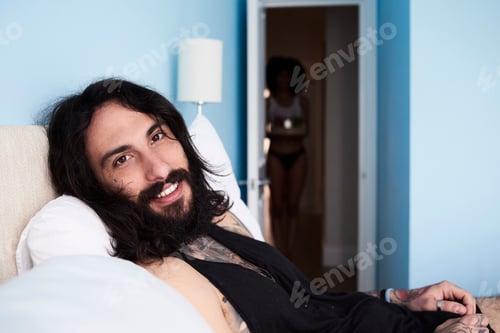 Preview: Portrait of smiling tattooed man lying n bed with girlfriend in background