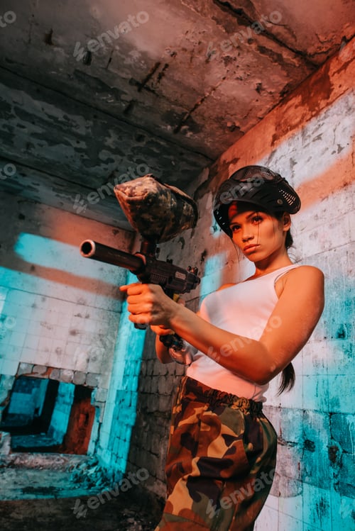 Preview: attractive young female paintballer in goggle mask above head aiming by paintball marker in