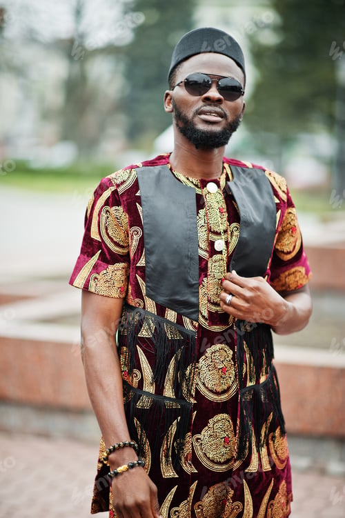 Preview: African stylish and handsome man in traditional outfit and black cap standing outdoor.