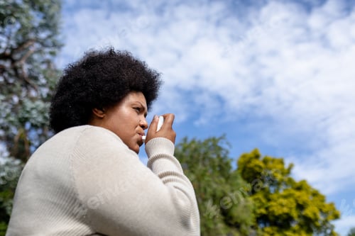 Preview: Low angle view of african american mid adult woman using asthma inhaler while standing against sky