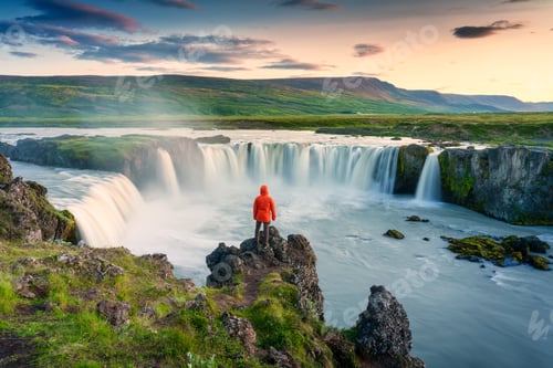 Preview: Godafoss waterfall flowing with colorful sunset sky and male tourist standing on cliff