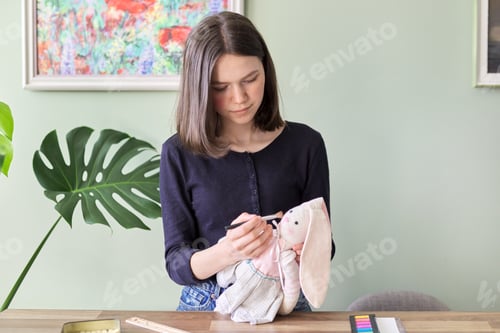Preview: Teenager girl sewing bunny doll toy, sitting at home at table, draws pink cheeks with eyeshadow