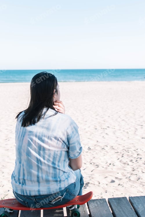 Preview: Teen girl in a tie dye shirt sitting on a skateboard looking at the beach