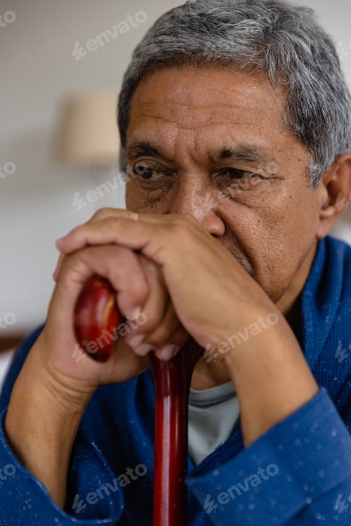Preview: Thoughtful senior biracial man sitting and holding walking stick at home