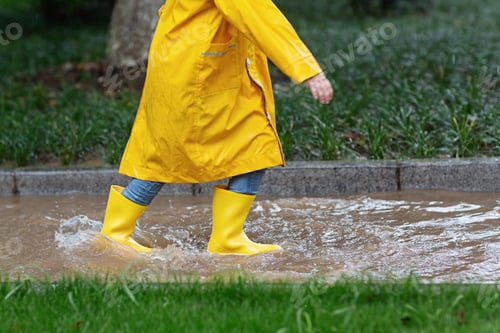 Preview: Kid in yellow raincoat and rubber boots jumping on puddle in park during rain at summer