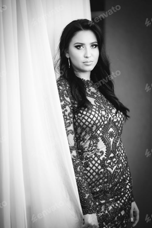 Preview: Vertical grayscale shot of a young Caucasian lady in an elegant dress and look near curtains