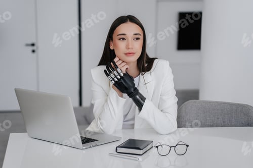 Preview: woman in white suit with artificial arm prosthesis sitting at desk with laptop looks aside dreamily