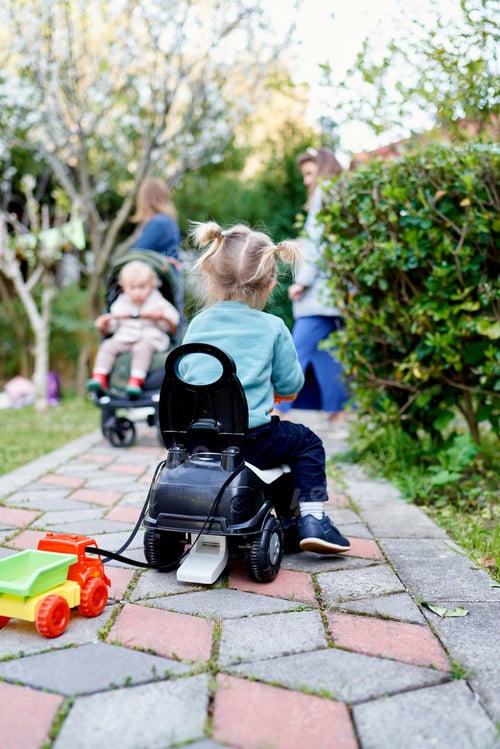 Preview: Little girl drives a toy car along a path in the garden with a toy truck tied to the seat. Back view