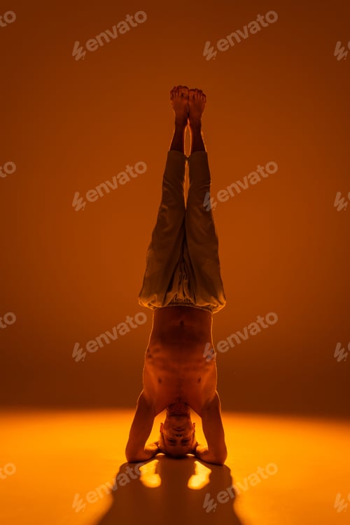 Preview: full length of shirtless man doing supported headstand yoga pose on brown