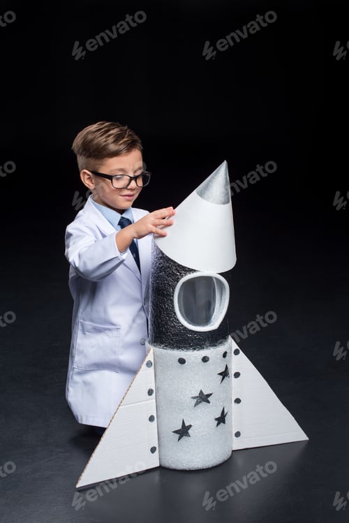 Preview: Smiling little boy in white coat and eyeglasses playing with toy rocket