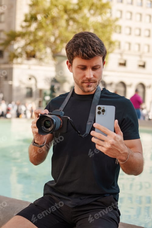Preview: Focused man using photo camera and smartphone near fountain on square