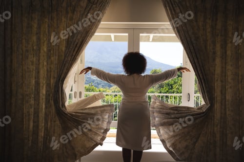 Preview: African american woman standing in front of window with sunny mountain view drawing curtains