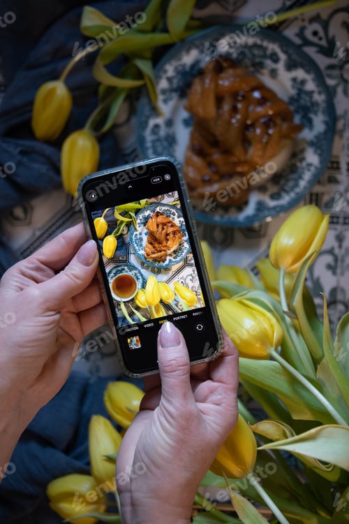 Preview: A woman takes pictures on a mobile phone of a table, sweet buns,a tea,yellow tulips,colorful flatley