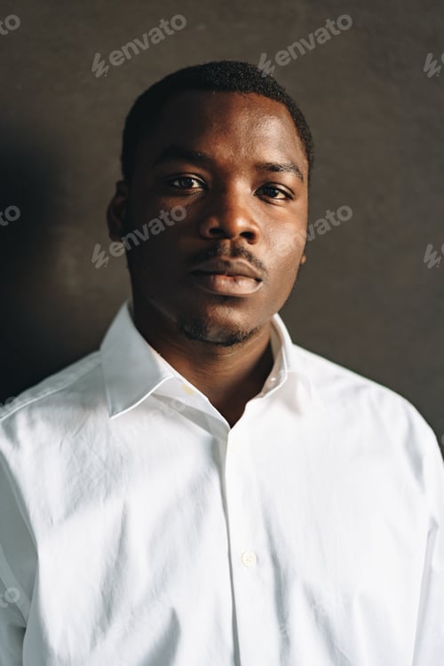 Preview: Close up portrait of young african man in white shirt against black wall in studio