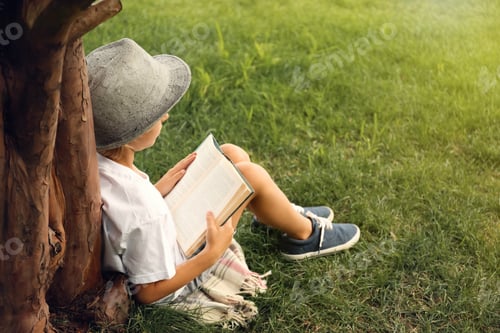 Preview: Cute little boy reading book on green grass near tree in park