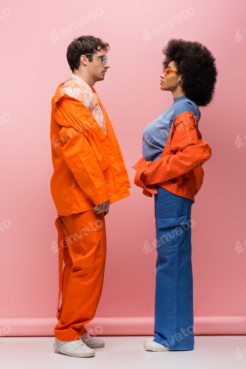 Preview: Side view of trendy interracial couple in sunglasses looking at each other on pink background