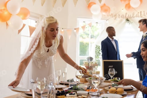 Preview: Bride arranging table setting at wedding reception with guests enjoying celebration
