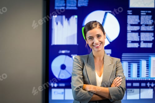 Preview: Woman standing arms crossed and smiling in office, digital screen showing pie charts and bar graphs