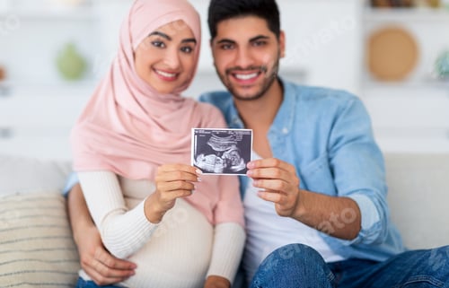 Preview: Happy arab family demonstrating ultrasound scan and smiling to camera, sitting on sofa at home