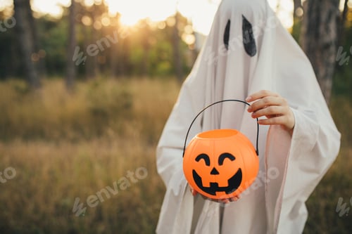 Preview: Girl wearing ghost costume holding pumpkin bucket with candies, standing in a forest.