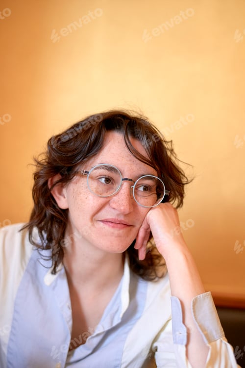 Preview: Smiling woman with glasses resting her chin on her hand against a warm background.