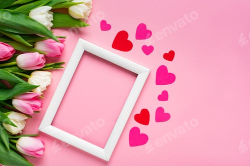 Preview: Top view of white frame with paper hearts and bouquet of tulips on pink surface
